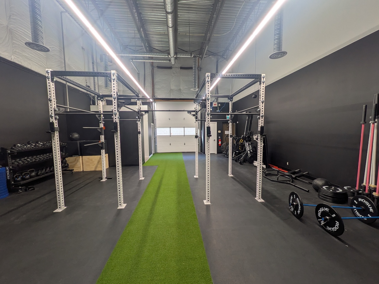 The Health Garage training gym: two white squat rigs either side of a green turf lane, with kettlebells, dumbbells and Rogue barbells along the walls.